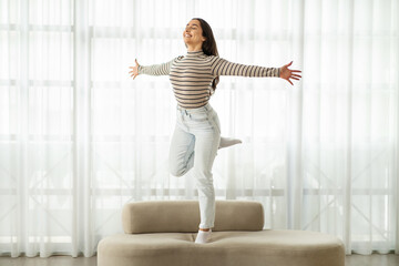 Exuberant young woman with a beaming smile leaps joyfully on a couch, arms outstretched
