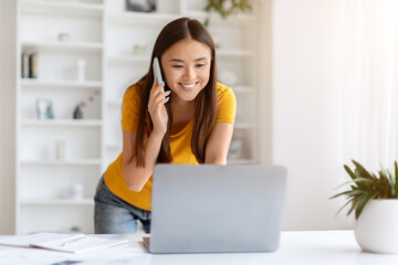 Young Asian Woman With Cellphone And Laptop Working Remotely From Home Office