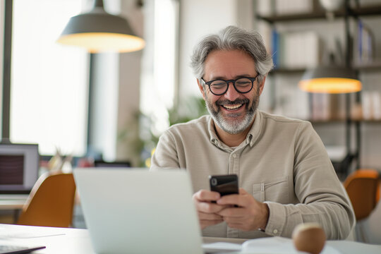 The image shows a smiling man with gray hair and glasses, looking at his smartphone in a bright office, with a laptop open in front of him and a blurred office background.