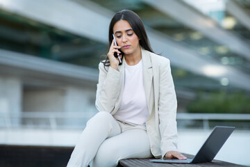 Upset arabic businesswoman talking on phone sitting with laptop outside