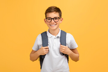 Cheerful schoolboy wearing glasses and backpack standing confidently on yellow studio background - obrazy, fototapety, plakaty
