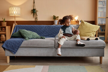 Full shot of African American girl child with ponytails sitting on big couch in cozy living room
