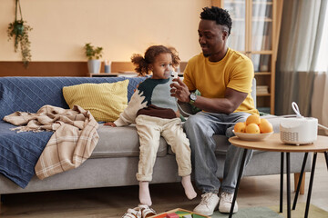 Obraz premium Full shot of smiling African American dad helping ill little daughter with inhalation through nebulizer while sitting on big sofa at home