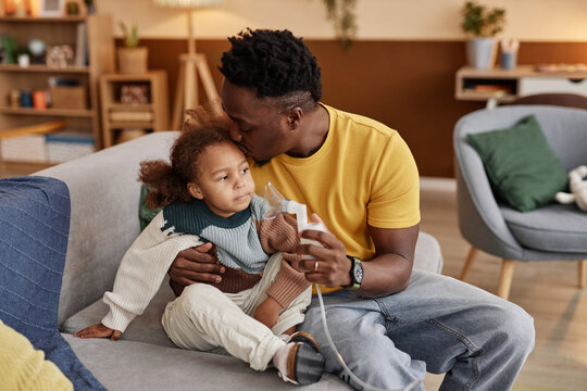Medium full shot of African American father kissing daughters forehead while sitting on couch with nebulizer mask in hand