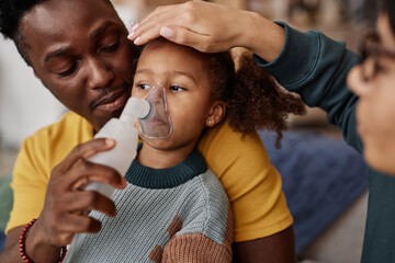 Medium close up shot of Black father with unwell little daughter holding nebulizer mask on her face while mother checking temperature putting hand on forehead