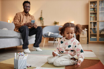 Full shot with focus on little African American girl in heart print sweatshirt sitting on living room floor holding watercolor marker, defocused smiling father sitting on couch watching her