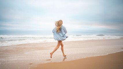 Fototapeta na wymiar Full length of carefree woman with blond hair running along sea on sandy beach. Rear view of female tourist in denim shirt on summer vacation. Excited lady is on beach holiday.