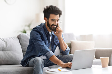 Smiling indian man multitasking, talking on phone while typing on laptop