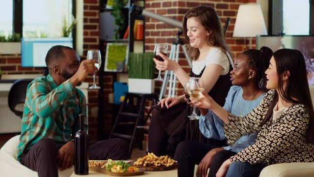 Friends laughing, toasting with alcoholic drinks, celebrating special occasion in cozy apartment living room. Colleagues spending time with each other during festivity at home