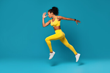 Fototapeta na wymiar Athletic black woman leaping during dynamic workout on blue backdrop