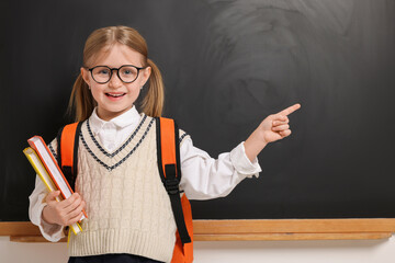 Happy schoolgirl in glasses with backpack and books pointing at something near blackboard, space for text