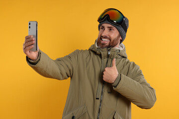 Winter sports. Happy man in ski suit and goggles taking selfie on orange background