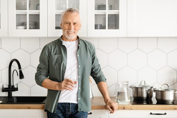 Cheerful mature man holding glass of water while posing in modern kitchen