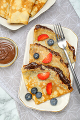 Tasty crepes with chocolate paste and berries served on white marble table, flat lay