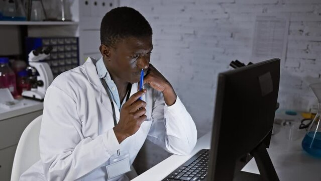 A thoughtful african man in a lab coat analyzing data on a computer in a laboratory setting.