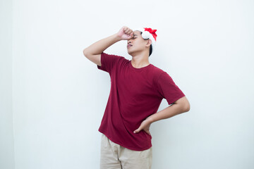 Fototapeta na wymiar Young Asian man wearing a Santa Claus hat expressing a headache by holding his head and showing signs of pain isolated by a white background for visual communication