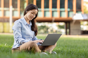 Content young woman in a blue shirt and brown pants enjoys working on her laptop while sitting...