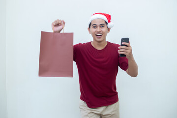 Young Asian man wearing a Santa Claus hat holding a smartphone and a shopping bag with expressions of smile, shock, and surprise, isolated against a white background for visual communication