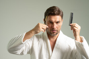 sexy man in bathrobe holding scissors and comb in front of mirror, looking in mirror, enjoying beauty care activity, satisfied with haircare cosmetic products, haircut, barber work result