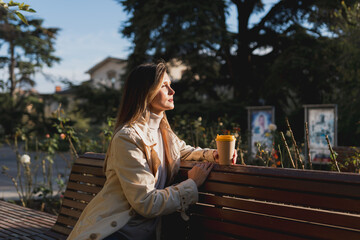 Fototapeta premium Woman drinks from cup on wooden bench. She is wearing a white shirt enjoying her beverage. The bench is located in a park setting, with trees in the background.