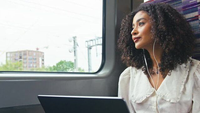 Young businesswoman commuting sitting on moving train passing through city working on laptop and listening to music or podcast on earphones looking out of window - shot in slow motion