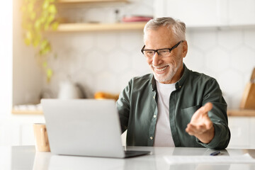 Cheerful elderly man wearing glasses making video call on laptop in kitchen