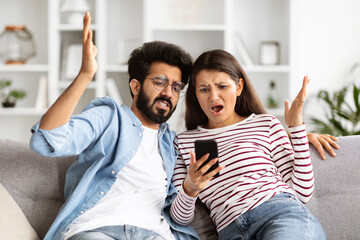 Furious indian couple looking at smartphone screen, home interior