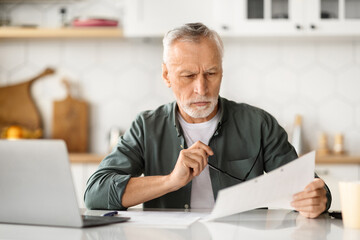 Pensive senior man holding paperwork, focused on reviewing documents in bright kitchen
