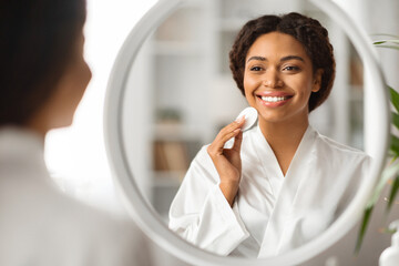 Beautiful black woman looking in mirror while cleansing skin with cotton pad - obrazy, fototapety, plakaty
