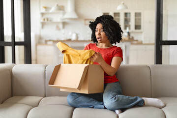 Surprised young black woman opening delivery box and looking at yellow t-shirt