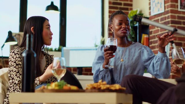 Diverse friends at house party paying attention to interesting story shared by woman. African american guest making asian person laugh in living room while enjoying drinks and charcuterie platter