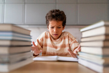 Puzzled black boy making homework surrounded by towering piles of books