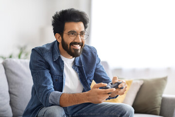 Cheerful young Indian man enjoying playing video games at home - obrazy, fototapety, plakaty
