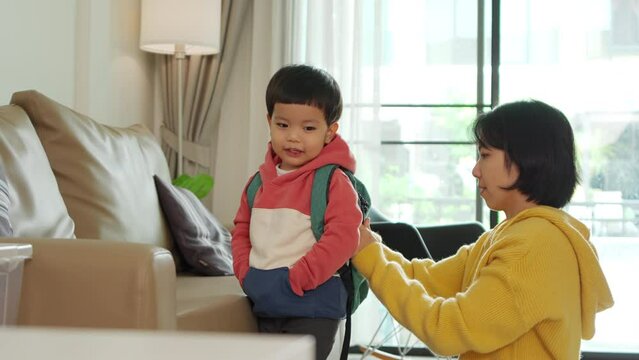 Asian Chinese mother helping little son to put a book into his backpack in Living room while preparing to school. Boy with a schoolbag