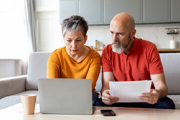Senior Spouses Reading Papers And Using Laptop Computer At Home