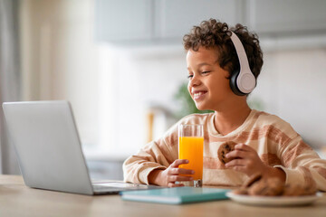 Black boy wearing headphones eating snacks while having online class on laptop