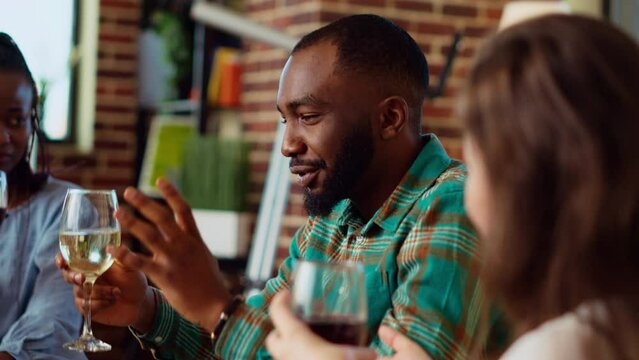 African american man entertaining friends at house party, making them pay attention to interesting story. Guest making BIPOC women laugh in living room while enjoying drinks and charcuterie platter