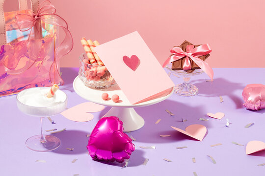 Cards, gift bags, heart balloons, paper hearts, chocolates, wafer rolls and candy balls are displayed on a pastel purple background. Valentine's Day party table.