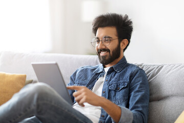 Naklejka premium Handsome young indian man with digital tablet relaxing on couch at home