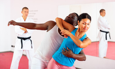 African-american man and Asian woman exercising elbow strike together in gym, their self-defence trainer in kimono standing and watching.