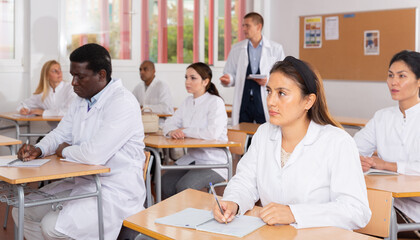 International group of different age people in white coats sitting in lecture hall at refresher...