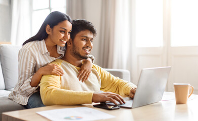 Happy indian couple websurfing on laptop together while relaxing at home