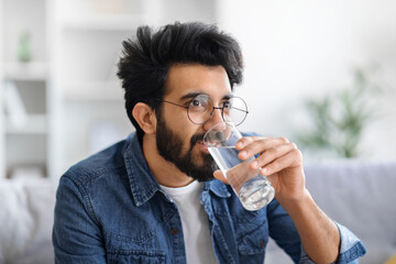 Young indian man wearing eyeglasses drinking water from clear glass at home