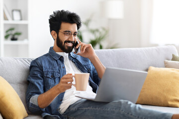 Relaxed indian man talking on cellphone, holding coffee and working on laptop