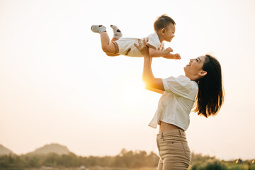 A woman holds her newborn baby up high, as they enjoy a moment of nature in the park at sunset. The...