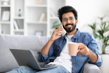 Cheerful indian man holding coffee cup, talking on cellphone and using laptop