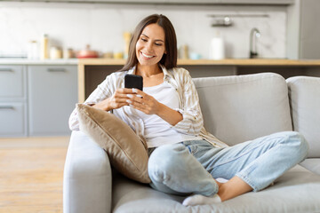 Cheerful young woman texting on her smartphone, sitting cross-legged on comfortable couch