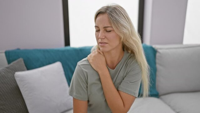A young woman in a casual t-shirt showing discomfort or neck pain inside a modern living room setting.