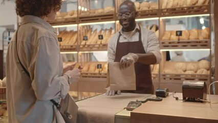 Medium shot of Caucasian female customer paying with card at bakery and Black salesman at counter smiling and giving her craft paper bag with takeout order