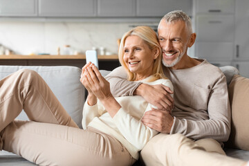 Happy senior couple sitting on sofa at home, using smartphone